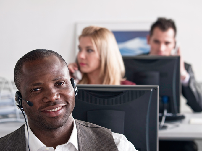 Young man sitting behind a desk in the customer support center and talking on the phone, in the blurred background other office workers are sitting and working.