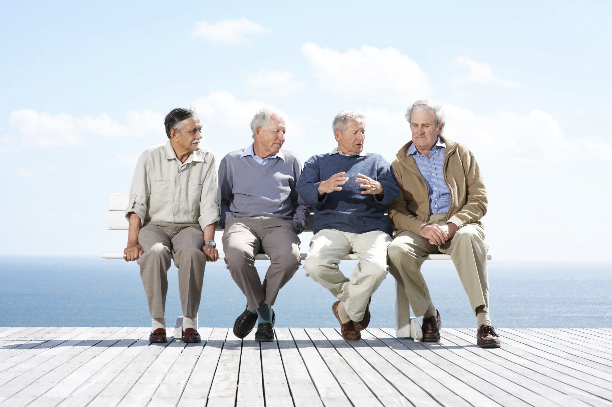 Four elderly men talking while sitting on a bench on a deck by the ocean