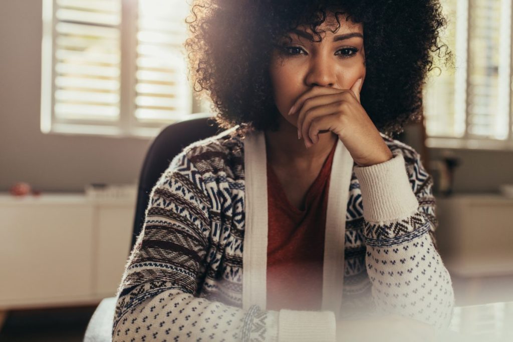 Female mental health clinician in a cardigan sitting at her desk, elbows on the table and hand over her mouth, reflecting on her competence and experiencing clinician imposter syndrome.