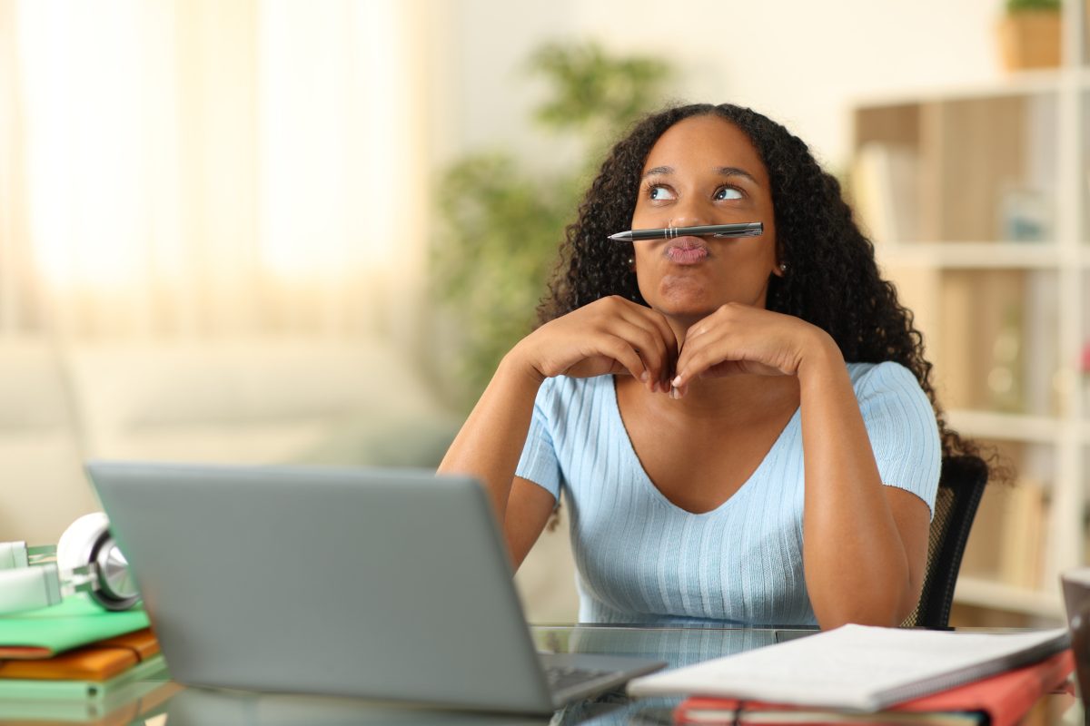 A young woman sits at a desk with a pencil balanced under her nose, gazing upward and appearing distracted while working on a laptop — illustrating signs of undiagnosed ADHD in adults.