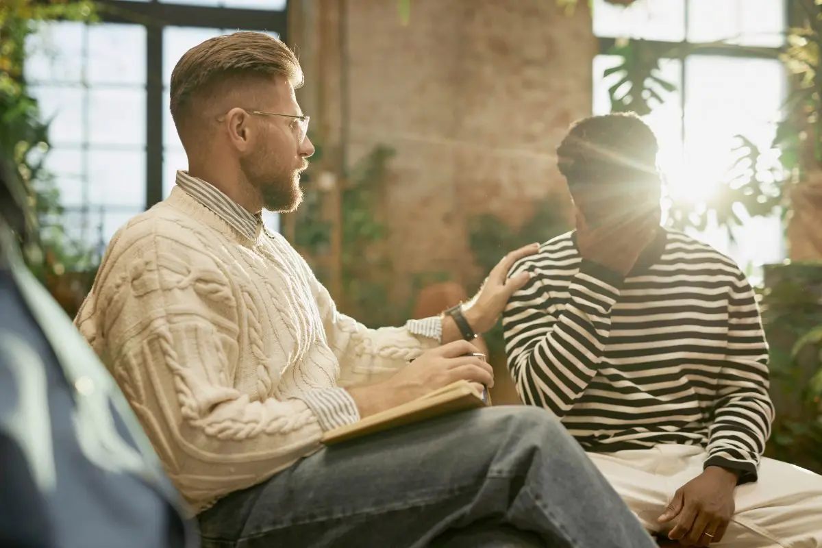 A psychologist holding a notepad places a supportive hand on a client's shoulder during an in-person behavioral health therapy session in a warm, plant-filled office setting