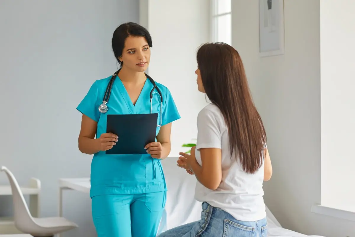 A female psychiatric nurse practitioner in scrubs with a stethoscope holds a clipboard while consulting with a female patient seated on an exam table in a behavioral health clinic