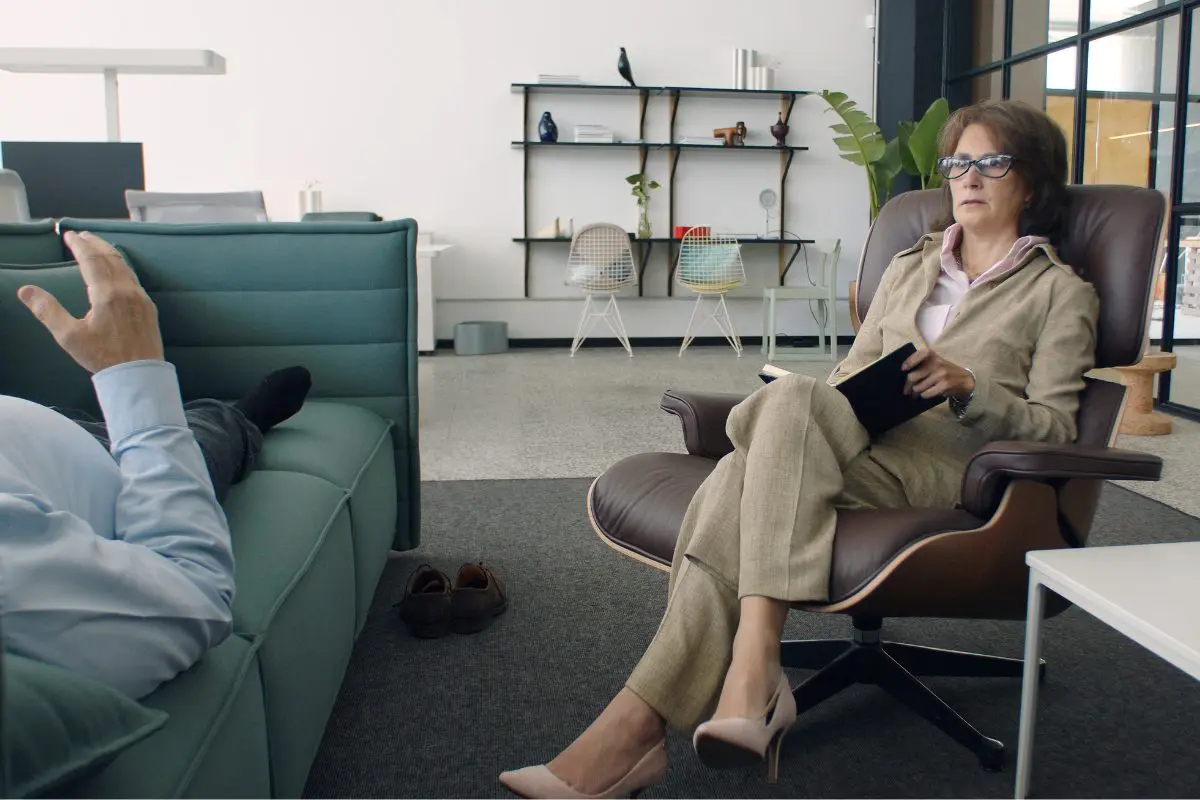 A female psychiatrist holding a notepad listens attentively to a patient reclining on a couch during an in-person psychiatric evaluation session in a professional office setting