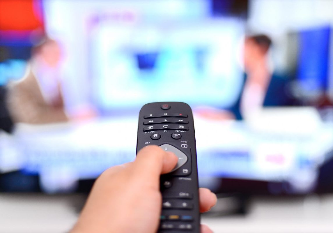 Image of political obsession disorder triggered by constant news exposure on TV Hand holding a TV remote pointed at political news broadcast, symbolizing political obsession disorder